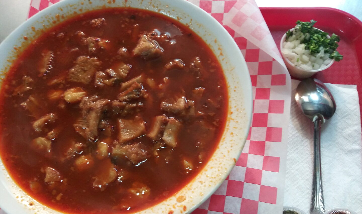 A bowl of menudo with onion, cilantro and a spoon on a red tray