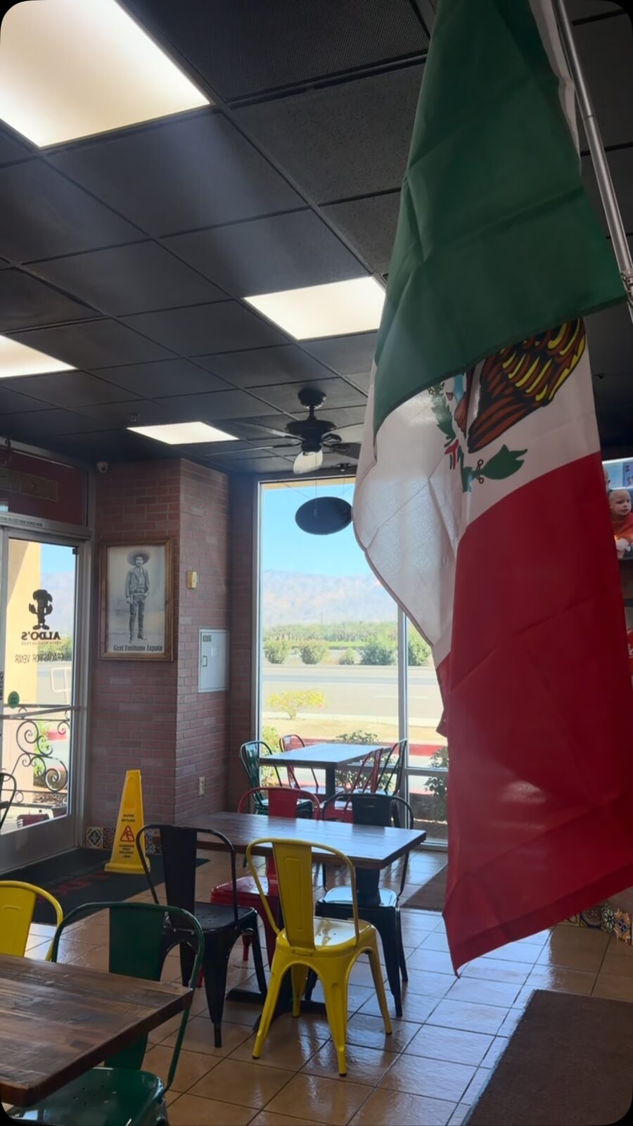 Inside Aldo's: tables, yellow chairs and a Mexican flag by the window